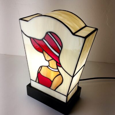 Lampe de table en vitrail de style Art Déco/Tiffany, représentant le profil d'une femme portant un large chapeau à bords. L'abat-jour trapézoïdal est principalement en verre ivoire opalescent. La figure féminine est réalisée avec du verre rouge vif pour la robe et le chapeau (avec des rayures blanches), et du verre ambre pour la peau. Le tout est serti de lignes de plomb noires. La lampe est posée sur un socle noir, et la lumière intérieure met en valeur le dessin graphique typique de l'Art Déco