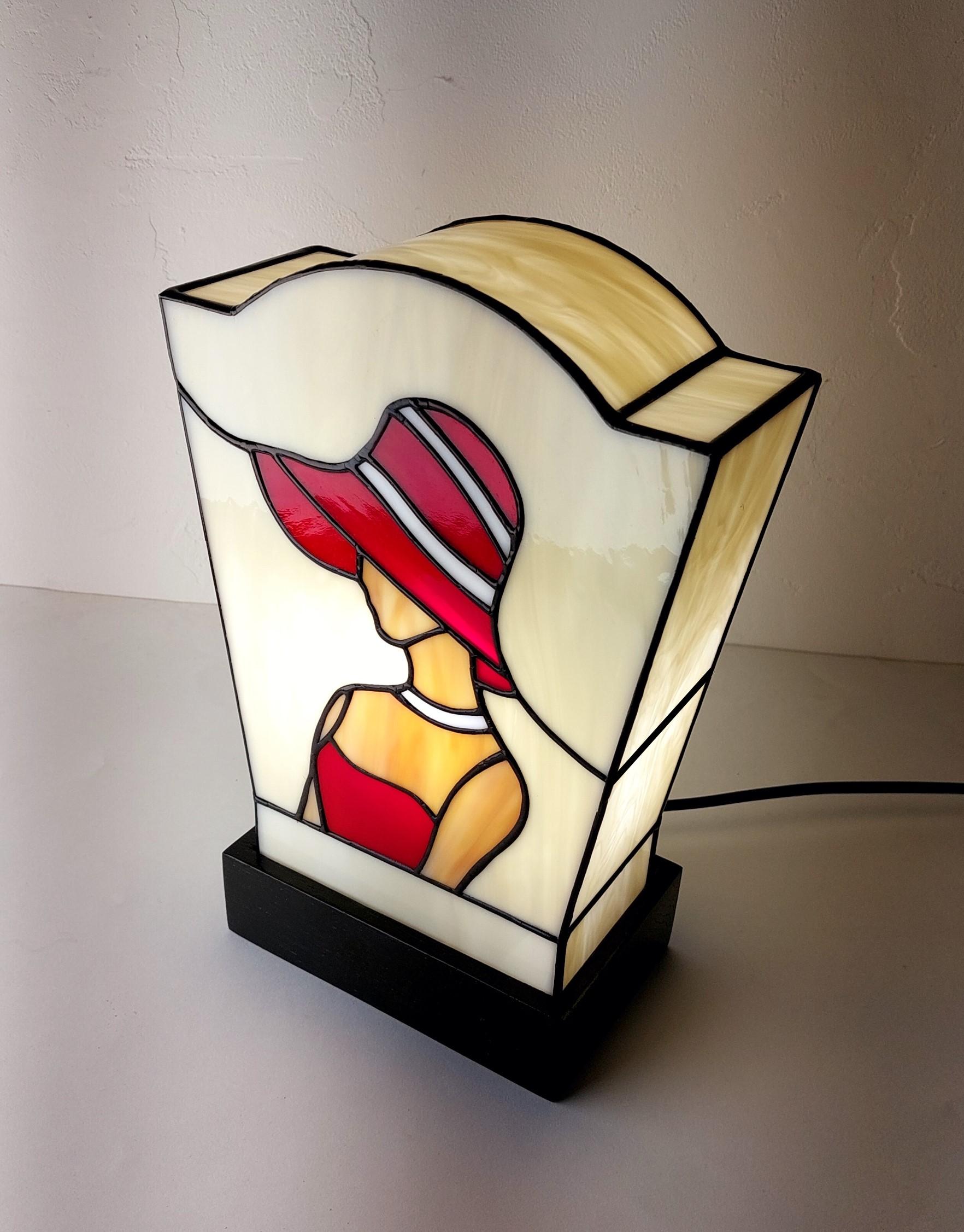 Lampe de table en vitrail de style Art Déco/Tiffany, représentant le profil d'une femme portant un large chapeau à bords. L'abat-jour trapézoïdal est principalement en verre ivoire opalescent. La figure féminine est réalisée avec du verre rouge vif pour la robe et le chapeau (avec des rayures blanches), et du verre ambre pour la peau. Le tout est serti de lignes de plomb noires. La lampe est posée sur un socle noir, et la lumière intérieure met en valeur le dessin graphique typique de l'Art Déco