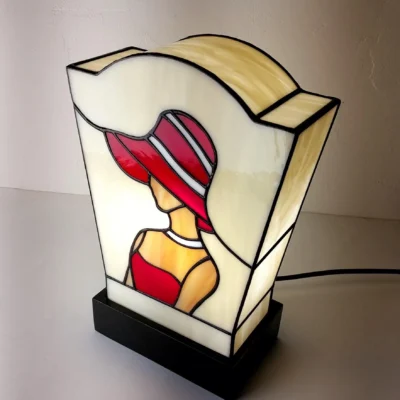 Lampe de table en vitrail de style Art Déco/Tiffany, représentant le profil d'une femme portant un large chapeau à bords. L'abat-jour trapézoïdal est principalement en verre ivoire opalescent. La figure féminine est réalisée avec du verre rouge vif pour la robe et le chapeau (avec des rayures blanches), et du verre ambre pour la peau. Le tout est serti de lignes de plomb noires. La lampe est posée sur un socle noir, et la lumière intérieure met en valeur le dessin graphique typique de l'Art Déco