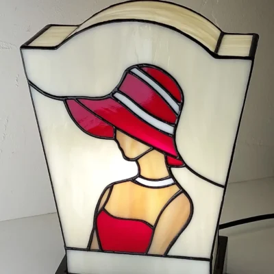 Lampe de table en vitrail de style Art Déco/Tiffany, représentant le profil d'une femme portant un large chapeau à bords. L'abat-jour trapézoïdal est principalement en verre ivoire opalescent. La figure féminine est réalisée avec du verre rouge vif pour la robe et le chapeau (avec des rayures blanches), et du verre ambre pour la peau. Le tout est serti de lignes de plomb noires. La lampe est posée sur un socle noir, et la lumière intérieure met en valeur le dessin graphique typique de l'Art Déco