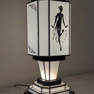 Lampe de table colonne Art Déco en vitrail Tiffany, de forme rectangulaire avec une base et un sommet étagés. L'abat-jour est bicolore, noir et blanc. Un panneau met en vedette une silhouette d'une danseuse de Charleston stylisée (Flapper) avec des perles et une jupe à franges, réalisée en verre noir opaque sur fond blanc opalescent. Les autres panneaux et la base présentent des motifs géométriques noirs et blancs. La lampe diffuse une lumière chaude, mettant en valeur son thème emblématique des années folles.