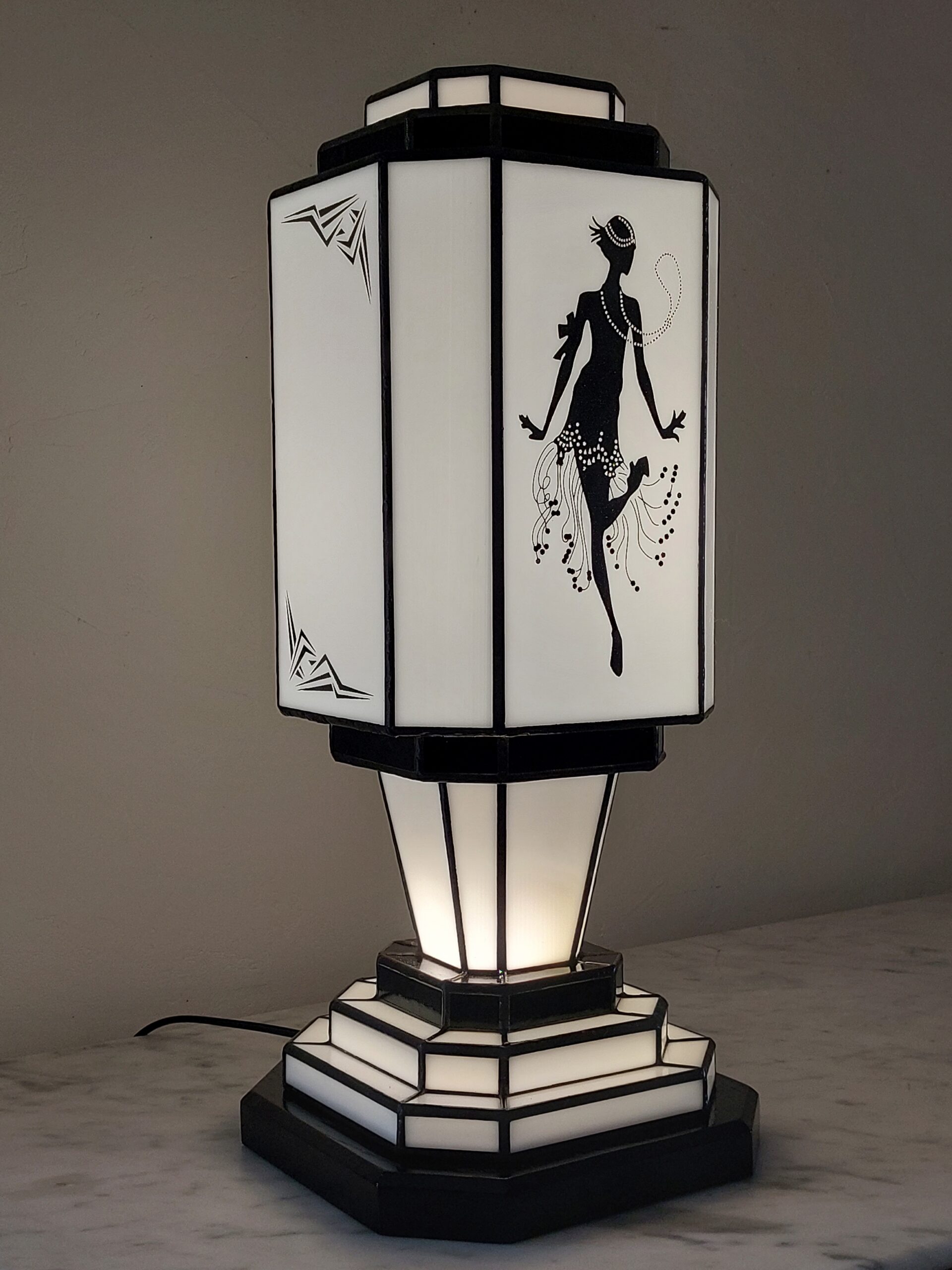 Lampe de table colonne Art Déco en vitrail Tiffany, de forme rectangulaire avec une base et un sommet étagés. L'abat-jour est bicolore, noir et blanc. Un panneau met en vedette une silhouette d'une danseuse de Charleston stylisée (Flapper) avec des perles et une jupe à franges, réalisée en verre noir opaque sur fond blanc opalescent. Les autres panneaux et la base présentent des motifs géométriques noirs et blancs. La lampe diffuse une lumière chaude, mettant en valeur son thème emblématique des années folles.