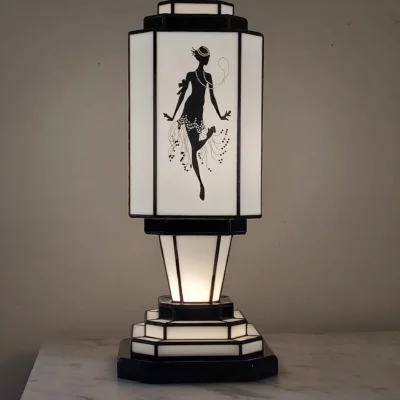 Lampe de table colonne Art Déco en vitrail Tiffany, de forme rectangulaire avec une base et un sommet étagés. L'abat-jour est bicolore, noir et blanc. Un panneau met en vedette une silhouette d'une danseuse de Charleston stylisée (Flapper) avec des perles et une jupe à franges, réalisée en verre noir opaque sur fond blanc opalescent. Les autres panneaux et la base présentent des motifs géométriques noirs et blancs. La lampe diffuse une lumière chaude, mettant en valeur son thème emblématique des années folles.