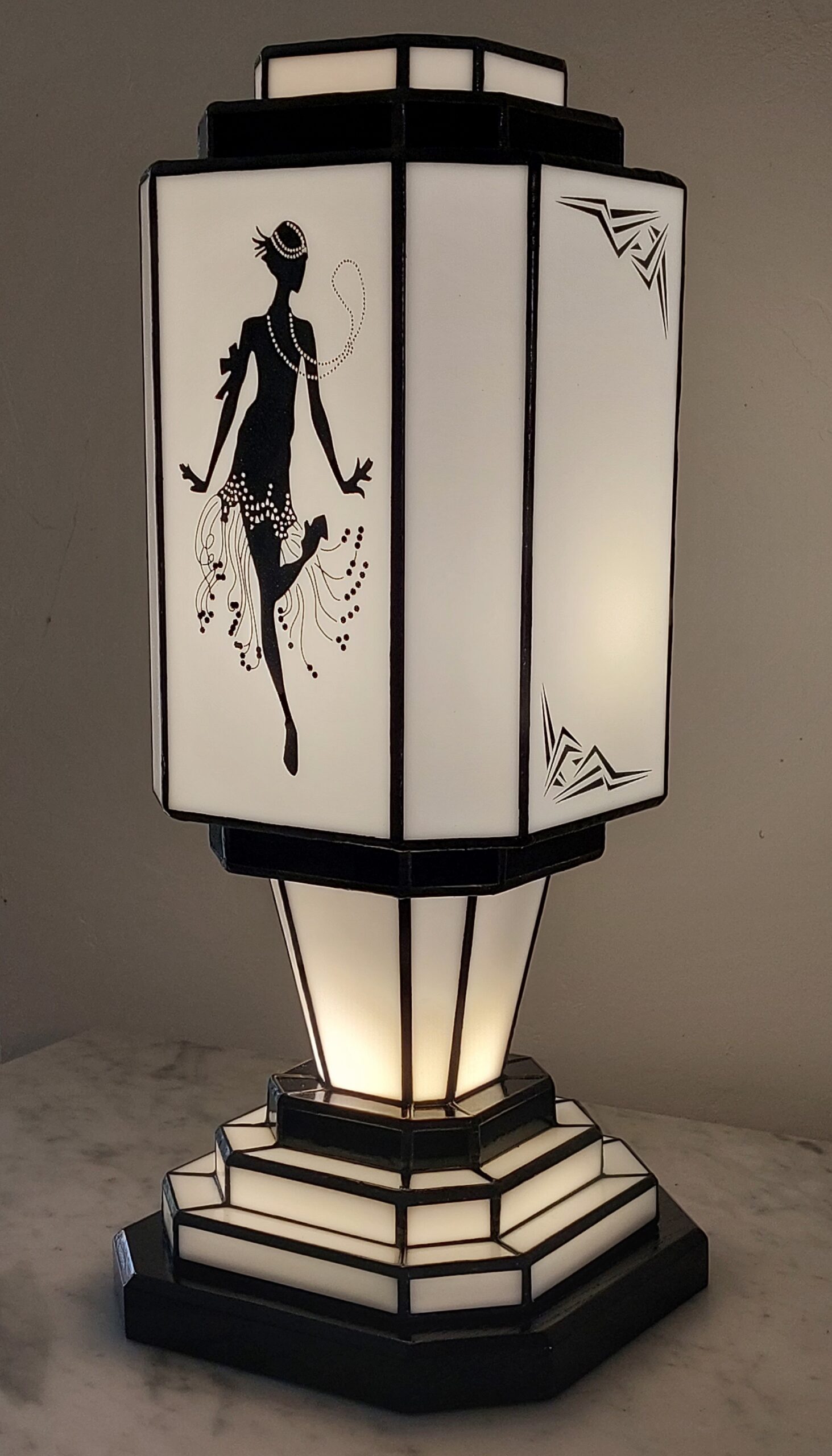 Lampe de table colonne Art Déco en vitrail Tiffany, de forme rectangulaire avec une base et un sommet étagés. L'abat-jour est bicolore, noir et blanc. Un panneau met en vedette une silhouette d'une danseuse de Charleston stylisée (Flapper) avec des perles et une jupe à franges, réalisée en verre noir opaque sur fond blanc opalescent. Les autres panneaux et la base présentent des motifs géométriques noirs et blancs. La lampe diffuse une lumière chaude, mettant en valeur son thème emblématique des années folles.