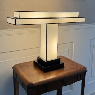 Lampe de table Art Déco en vitrail Tiffany, de forme rectangulaire et horizontale, rappelant un design Bauhaus ou une structure architecturale. La base est carrée et étagée, en noir. Un pilier vertical central en verre blanc dépoli soutient un abat-jour composé de deux longs rectangles horizontaux superposés, en verre blanc opalescent et noir opaque. Les soudures sont noires. La lampe est illuminée de l'intérieur, diffusant une lumière douce et homogène qui met en valeur sa géométrie épurée et son contraste noir et blanc, posée sur une petite table en bois vintage.