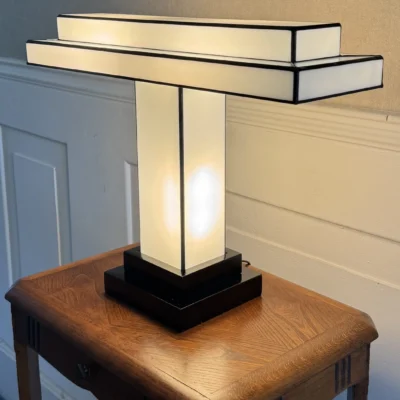 Lampe de table Art Déco en vitrail Tiffany, de forme rectangulaire et horizontale, rappelant un design Bauhaus ou une structure architecturale. La base est carrée et étagée, en noir. Un pilier vertical central en verre blanc dépoli soutient un abat-jour composé de deux longs rectangles horizontaux superposés, en verre blanc opalescent et noir opaque. Les soudures sont noires. La lampe est illuminée de l'intérieur, diffusant une lumière douce et homogène qui met en valeur sa géométrie épurée et son contraste noir et blanc, posée sur une petite table en bois vintage.