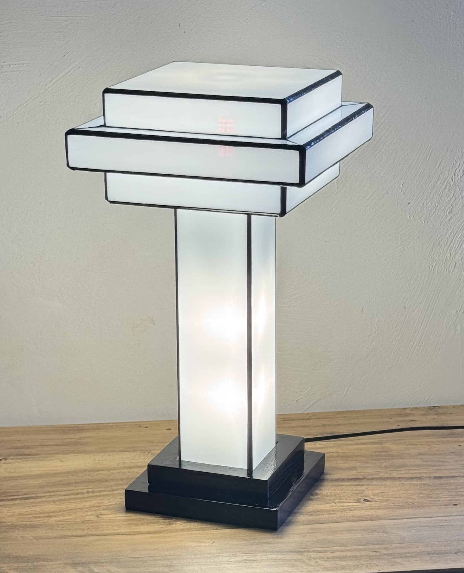 Lampe de table Art Déco en vitrail Tiffany, de forme cubique et étagée, évoquant un gratte-ciel stylisé. La base est carrée et noire. Un pilier vertical central en verre blanc dépoli s'élève vers un abat-jour composé de plusieurs plateaux carrés superposés, alternant entre le blanc opalescent et le noir opaque. Les soudures sont noires. La lampe est illuminée de l'intérieur, diffusant une lumière douce et homogène qui met en valeur sa géométrie architecturale et son contraste noir et blanc.