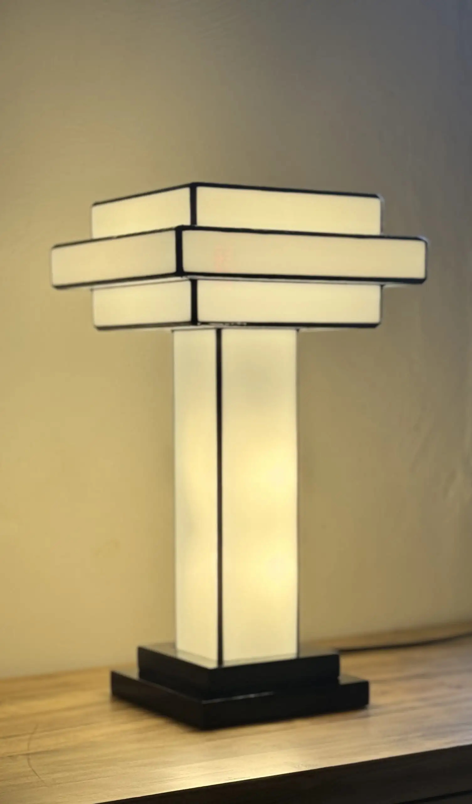 Lampe de table Art Déco en vitrail Tiffany, de forme cubique et étagée, évoquant un gratte-ciel stylisé. La base est carrée et noire. Un pilier vertical central en verre blanc dépoli s'élève vers un abat-jour composé de plusieurs plateaux carrés superposés, alternant entre le blanc opalescent et le noir opaque. Les soudures sont noires. La lampe est illuminée de l'intérieur, diffusant une lumière douce et homogène qui met en valeur sa géométrie architecturale et son contraste noir et blanc.