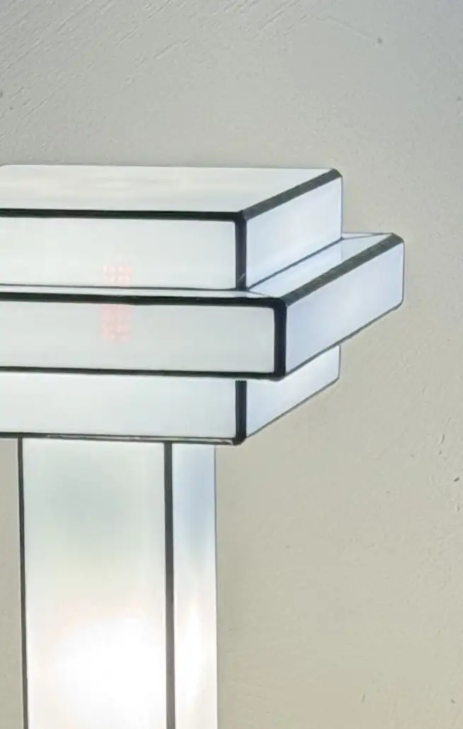 Lampe de table Art Déco en vitrail Tiffany, de forme cubique et étagée, évoquant un gratte-ciel stylisé. La base est carrée et noire. Un pilier vertical central en verre blanc dépoli s'élève vers un abat-jour composé de plusieurs plateaux carrés superposés, alternant entre le blanc opalescent et le noir opaque. Les soudures sont noires. La lampe est illuminée de l'intérieur, diffusant une lumière douce et homogène qui met en valeur sa géométrie architecturale et son contraste noir et blanc.