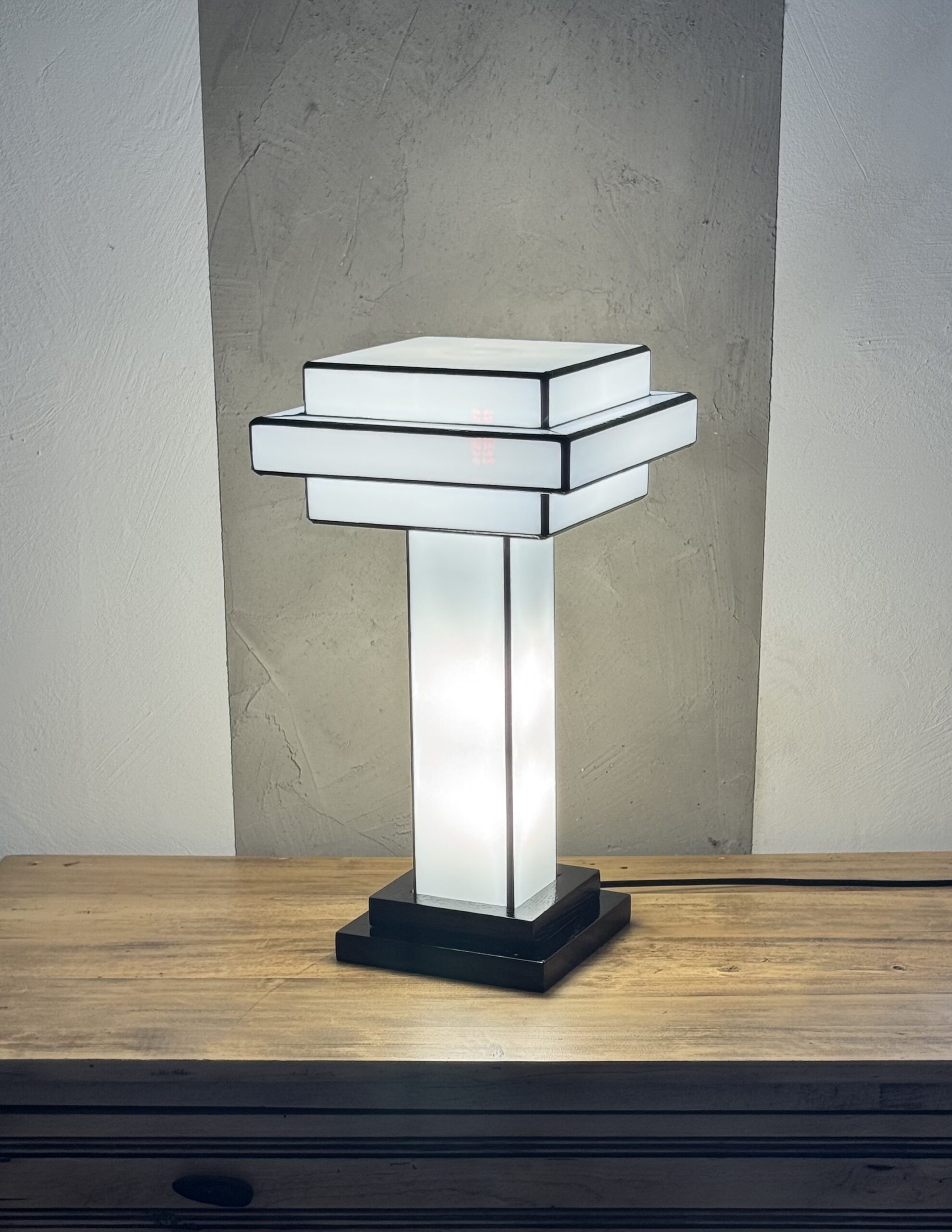 Lampe de table Art Déco en vitrail Tiffany, de forme cubique et étagée, évoquant un gratte-ciel stylisé. La base est carrée et noire. Un pilier vertical central en verre blanc dépoli s'élève vers un abat-jour composé de plusieurs plateaux carrés superposés, alternant entre le blanc opalescent et le noir opaque. Les soudures sont noires. La lampe est illuminée de l'intérieur, diffusant une lumière douce et homogène qui met en valeur sa géométrie architecturale et son contraste noir et blanc.