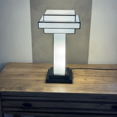 Lampe de table Art Déco en vitrail Tiffany, de forme cubique et étagée, évoquant un gratte-ciel stylisé. La base est carrée et noire. Un pilier vertical central en verre blanc dépoli s'élève vers un abat-jour composé de plusieurs plateaux carrés superposés, alternant entre le blanc opalescent et le noir opaque. Les soudures sont noires. La lampe est illuminée de l'intérieur, diffusant une lumière douce et homogène qui met en valeur sa géométrie architecturale et son contraste noir et blanc.