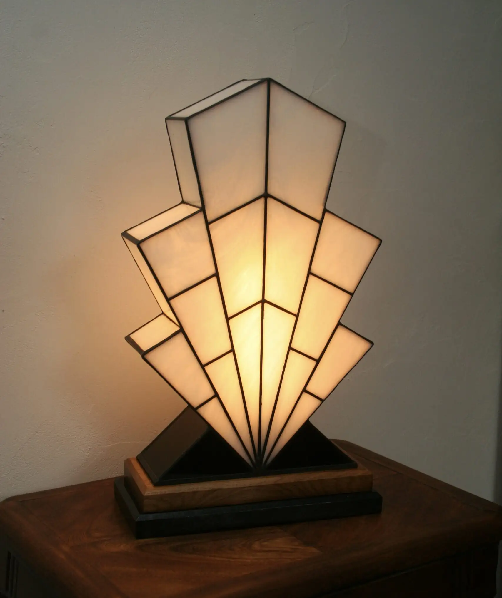 Grande lampe de table Art Déco en vitrail Tiffany, de forme spectaculaire et évasée, évoquant la silhouette d'un gratte-ciel ou un éventail stylisé ("Sunburst"). L'abat-jour est entièrement composé de panneaux de verre Blanc Opalescent, séparés par des soudures noires fines, créant un effet de rayons ou de marches. Elle repose sur une base étagée en bois foncé et noir mat, et diffuse une lumière blanche et chaude, mettant en évidence son architecture géométrique.