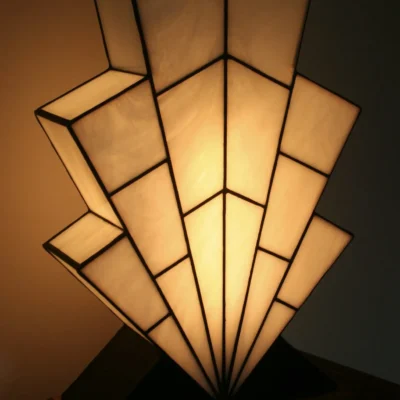 Grande lampe de table Art Déco en vitrail Tiffany, de forme spectaculaire et évasée, évoquant la silhouette d'un gratte-ciel ou un éventail stylisé ("Sunburst"). L'abat-jour est entièrement composé de panneaux de verre Blanc Opalescent, séparés par des soudures noires fines, créant un effet de rayons ou de marches. Elle repose sur une base étagée en bois foncé et noir mat, et diffuse une lumière blanche et chaude, mettant en évidence son architecture géométrique.