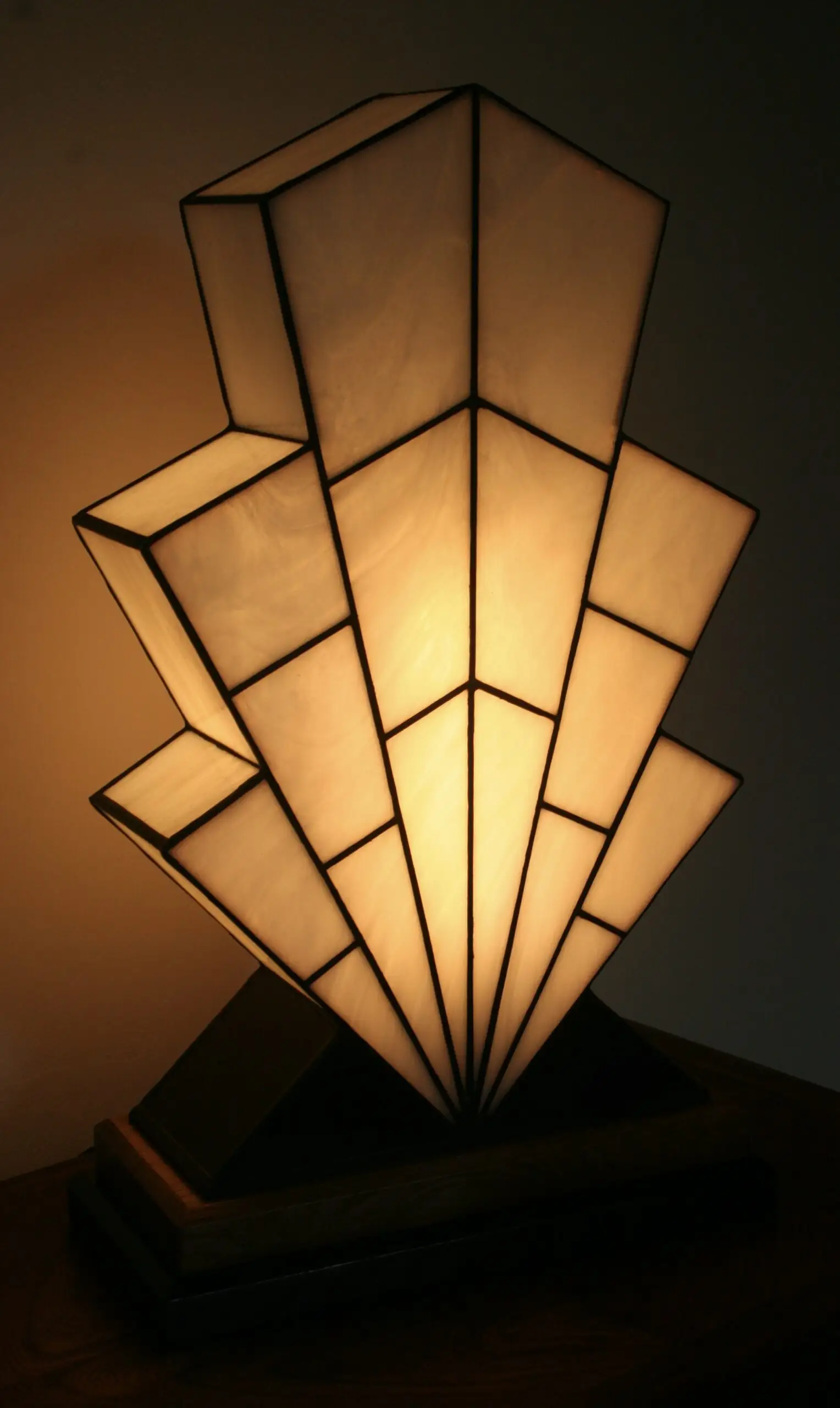 Grande lampe de table Art Déco en vitrail Tiffany, de forme spectaculaire et évasée, évoquant la silhouette d'un gratte-ciel ou un éventail stylisé ("Sunburst"). L'abat-jour est entièrement composé de panneaux de verre Blanc Opalescent, séparés par des soudures noires fines, créant un effet de rayons ou de marches. Elle repose sur une base étagée en bois foncé et noir mat, et diffuse une lumière blanche et chaude, mettant en évidence son architecture géométrique.