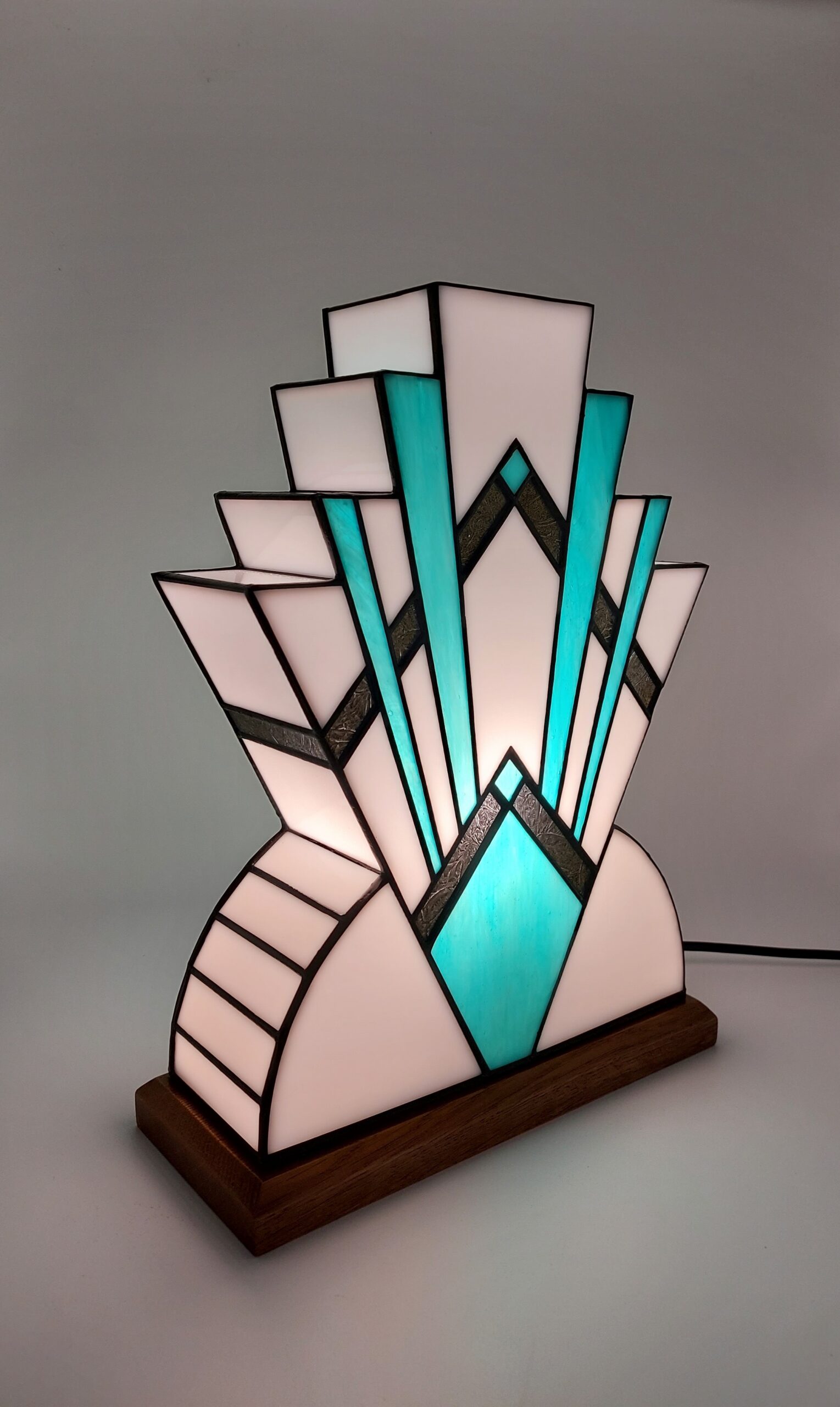 Lampe Art Déco en vitrail, inspiration Tiffany. Elle présente une base semi-circulaire blanche et un motif rayonnant et étagé de triangles vert-canard et blancs, rappelant un gratte-ciel ou un éventail stylisé, avec 2 bandes centrales dorées texturée.
