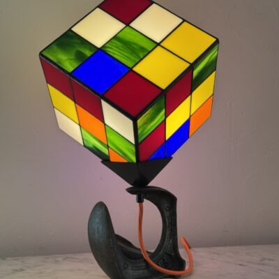 Lampe de table d'art moderne intitulée "Le Nostal Kub". L'abat-jour en forme de cube imite un Rubik's Cube non résolu, réalisé en vitrail de style Tiffany avec des carrés de verre coloré (rouge, jaune, bleu, vert, blanc cassé, orange). Le cube est monté sur un support industriel sombre qui ressemble à un sabot ou à un fer à cheval inversé. Un cordon électrique orange vif sort de la base. La lampe est posée sur une surface en marbre clair contre un mur blanc-gris.