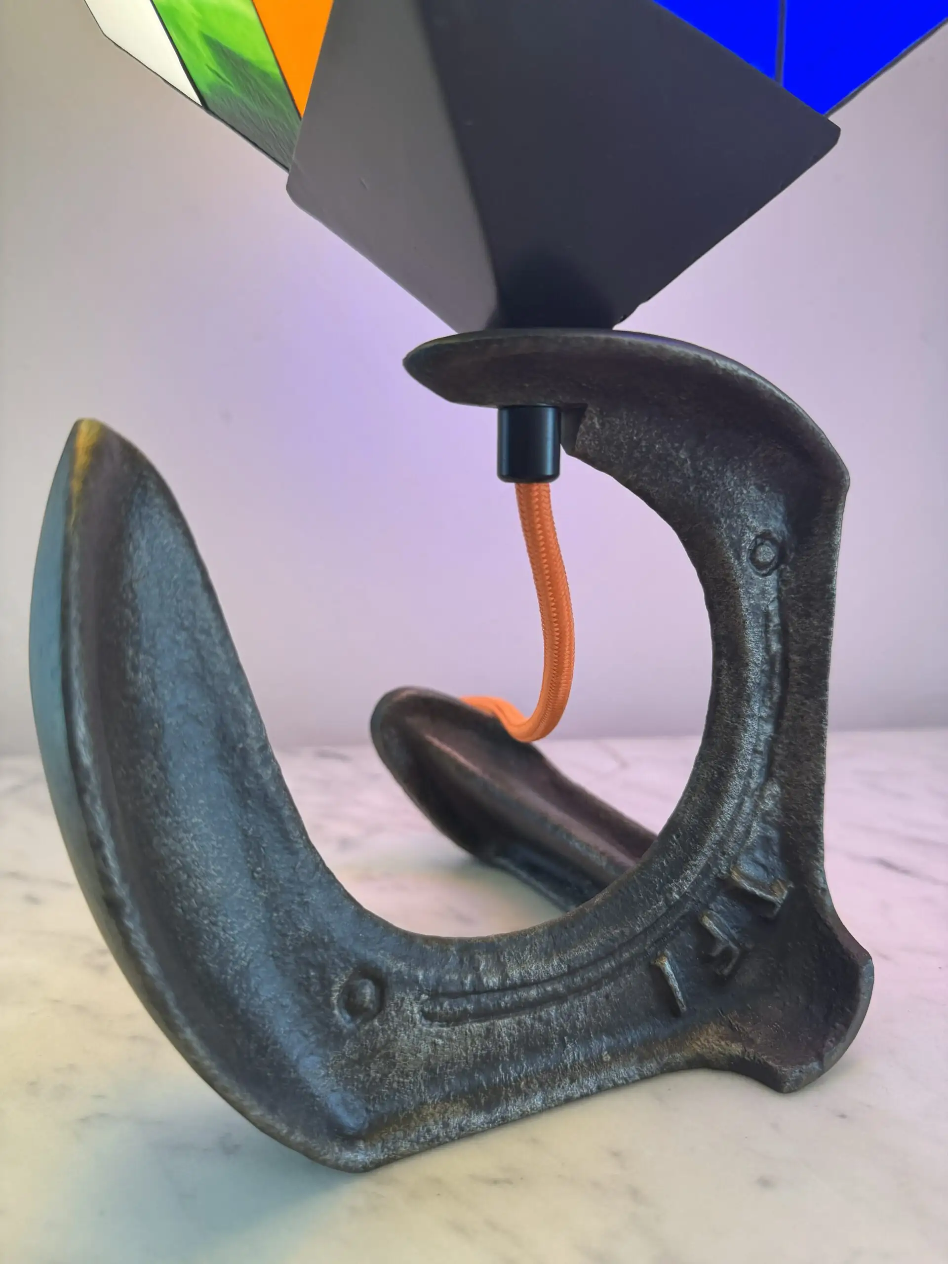 Lampe de table d'art moderne intitulée "Le Nostal Kub". L'abat-jour en forme de cube imite un Rubik's Cube non résolu, réalisé en vitrail de style Tiffany avec des carrés de verre coloré (rouge, jaune, bleu, vert, blanc cassé, orange). Le cube est monté sur un support industriel sombre qui ressemble à un sabot ou à un fer à cheval inversé. Un cordon électrique orange vif sort de la base. La lampe est posée sur une surface en marbre clair contre un mur blanc-gris.