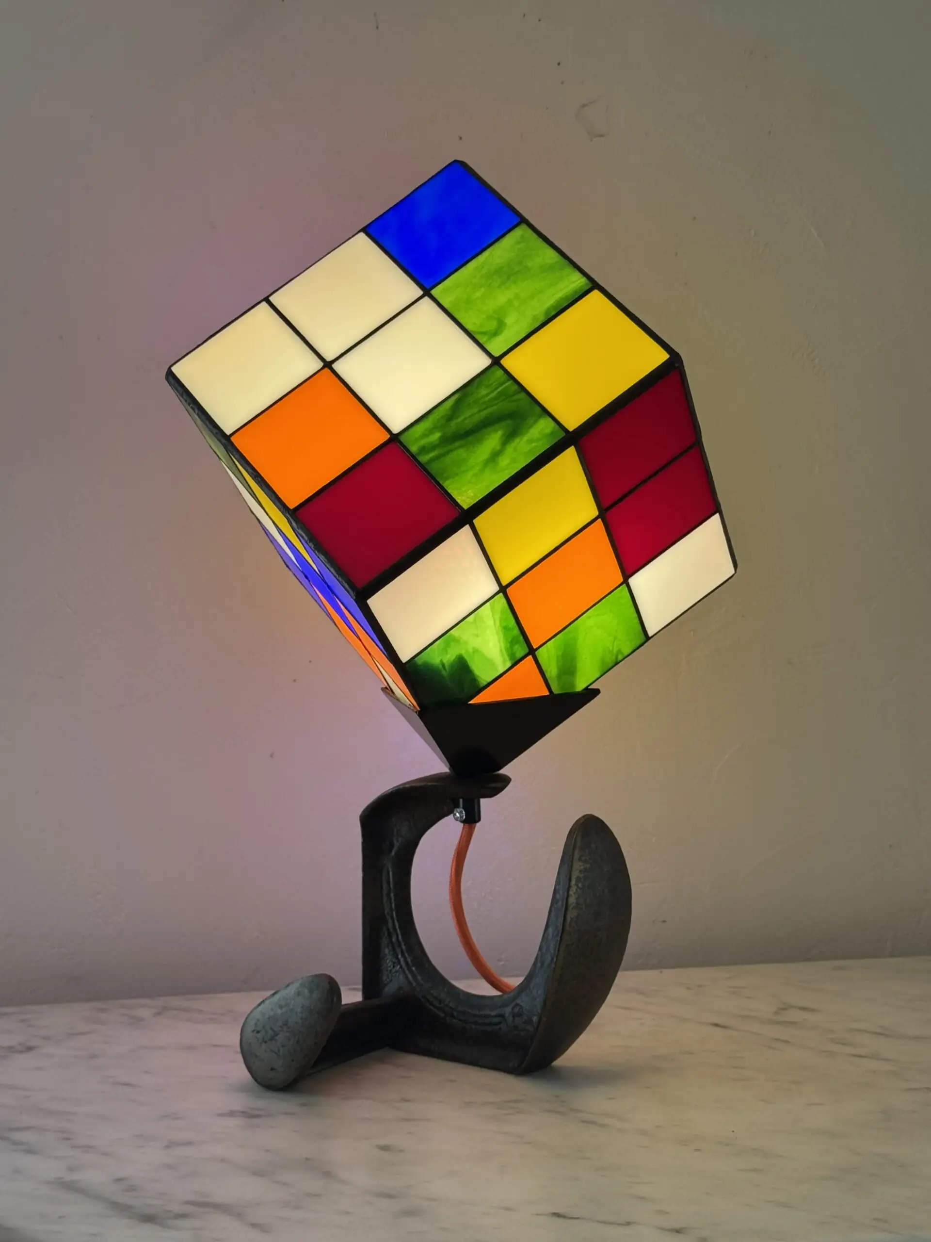 Lampe de table d'art moderne intitulée "Le Nostal Kub". L'abat-jour en forme de cube imite un Rubik's Cube non résolu, réalisé en vitrail de style Tiffany avec des carrés de verre coloré (rouge, jaune, bleu, vert, blanc cassé, orange). Le cube est monté sur un support industriel sombre qui ressemble à un sabot ou à un fer à cheval inversé. Un cordon électrique orange vif sort de la base. La lampe est posée sur une surface en marbre clair contre un mur blanc-gris.