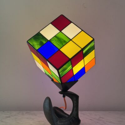 Lampe de table d'art moderne intitulée "Le Nostal Kub". L'abat-jour en forme de cube imite un Rubik's Cube non résolu, réalisé en vitrail de style Tiffany avec des carrés de verre coloré (rouge, jaune, bleu, vert, blanc cassé, orange). Le cube est monté sur un support industriel sombre qui ressemble à un sabot ou à un fer à cheval inversé. Un cordon électrique orange vif sort de la base. La lampe est posée sur une surface en marbre clair contre un mur blanc-gris.