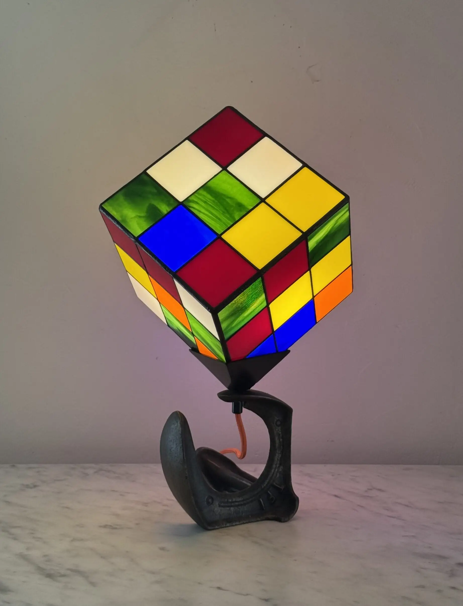 Lampe de table d'art moderne intitulée "Le Nostal Kub". L'abat-jour en forme de cube imite un Rubik's Cube non résolu, réalisé en vitrail de style Tiffany avec des carrés de verre coloré (rouge, jaune, bleu, vert, blanc cassé, orange). Le cube est monté sur un support industriel sombre qui ressemble à un sabot ou à un fer à cheval inversé. Un cordon électrique orange vif sort de la base. La lampe est posée sur une surface en marbre clair contre un mur blanc-gris.