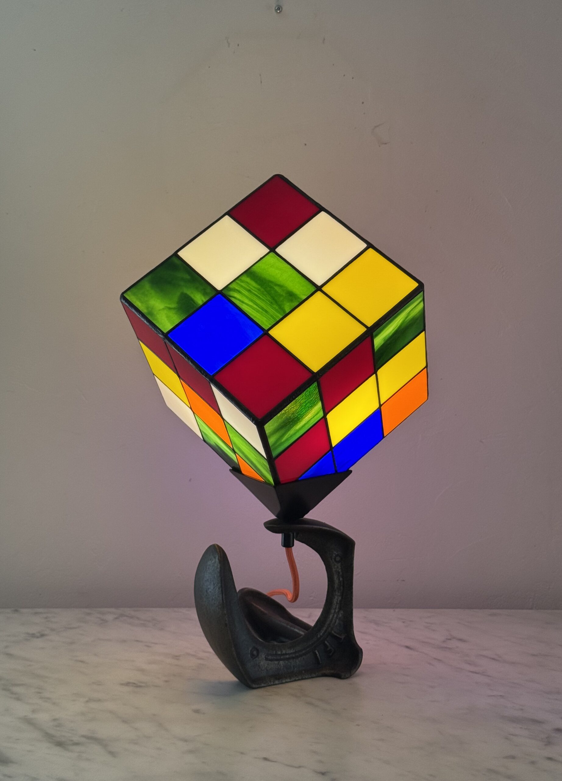 Lampe de table d'art moderne intitulée "Le Nostal Kub". L'abat-jour en forme de cube imite un Rubik's Cube non résolu, réalisé en vitrail de style Tiffany avec des carrés de verre coloré (rouge, jaune, bleu, vert, blanc cassé, orange). Le cube est monté sur un support industriel sombre qui ressemble à un sabot ou à un fer à cheval inversé. Un cordon électrique orange vif sort de la base. La lampe est posée sur une surface en marbre clair contre un mur blanc-gris.