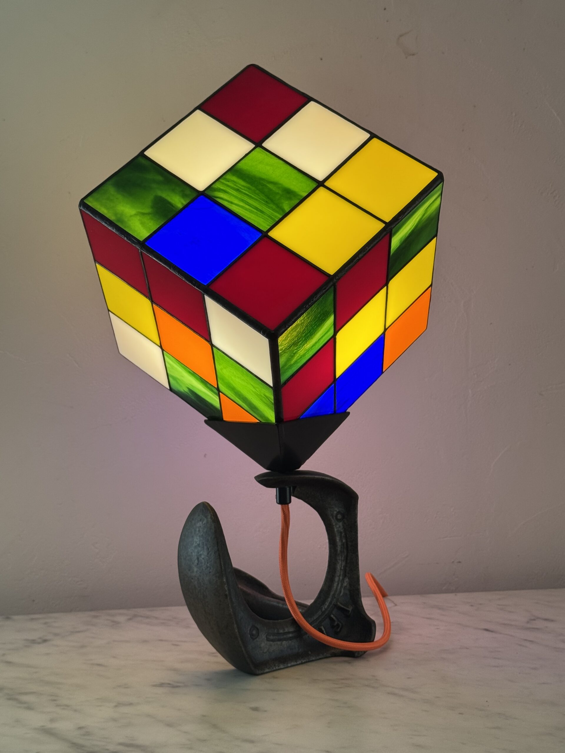 Lampe de table d'art moderne intitulée "Le Nostal Kub". L'abat-jour en forme de cube imite un Rubik's Cube non résolu, réalisé en vitrail de style Tiffany avec des carrés de verre coloré (rouge, jaune, bleu, vert, blanc cassé, orange). Le cube est monté sur un support industriel sombre qui ressemble à un sabot ou à un fer à cheval inversé. Un cordon électrique orange vif sort de la base. La lampe est posée sur une surface en marbre clair contre un mur blanc-gris.