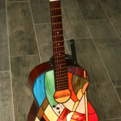 Lampe de sol artistique créée à partir d'une guitare acoustique transformée. La caisse de résonance de la guitare a été remplacée par un panneau en vitrail de style Tiffany, aux motifs abstraits et géométriques. Le vitrail intègre une palette de couleurs vives : bleu ciel, vert émeraude, rouge, orange, jaune, et verre irisé. Le corps en vitrail est éclairé par l'intérieur, illuminant le motif qui inclut de petits détails comme des notes de musique stylisées près de l'ouïe. La guitare est posée sur un support de sol noir sur un carrelage effet bois.