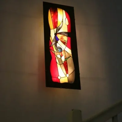 Très grande applique murale ou tableau lumineux rectangulaire, en vitrail Tiffany de style contemporain/abstrait, fixée sur un mur blanc. L'œuvre présente des formes géométriques et organiques entrelacées dans des teintes chaudes et vibrantes : rouge rubis, orange, jaune doré, avec des touches de bleu, de noir et de blanc. L'éclairage intense par l'arrière fait ressortir la richesse et la translucidité du verre coloré. Le panneau lumineux est orienté verticalement et est installé au-dessus d'une rampe d'escalier blanche
