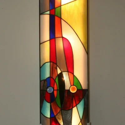 Très grande applique murale ou tableau lumineux vertical, en vitrail de style abstrait et contemporain, fixé sur un mur blanc. La composition est un assemblage de formes géométriques et courbes avec des lignes de plomb noires. Le panneau présente une riche palette de couleurs, incluant des bandes verticales rouges et bleues intenses, de grands segments jaunes et ambre, et des cercles concentriques bleus, rouges et oranges dans la partie inférieure. Un petit cercle jaune vif est visible en haut à gauche. L'éclairage par l'arrière révèle la texture et la translucidité du verre.
