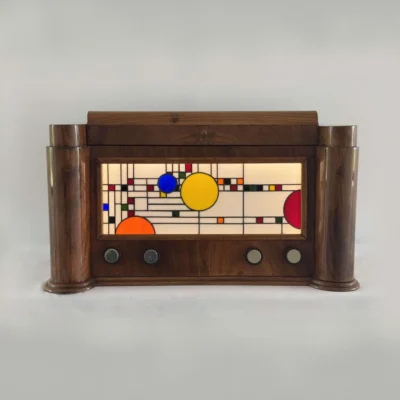 Coffre en bois foncé poli, de style Art Moderne, avec des formes arrondies sur les côtés. La façade principale est ornée d'un panneau de vitrail rétroéclairé. Le motif du vitrail est abstrait et géométrique, inspiré par le mouvement De Stijl (Piet Mondrian), composé de rectangles et de lignes noires sur fond blanc opalescent, intégrant des cercles colorés (jaune, bleu, orange, rouge). Le coffre possède deux tiroirs cachés sur les côtés et un couvercle supérieur qui s'ouvre pour révéler un miroir et un compartiment. Trois boutons ronds sont alignés sous le vitrail. L'éclairage LED interne diffuse une lumière douce à travers le vitrail.