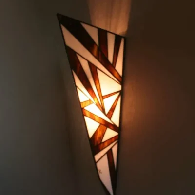 Applique murale verticale de style Art Déco en vitrail Tiffany, de forme effilée et conique, rappelant une pointe de flèche. Le motif est composé de multiples triangles et formes angulaires, créant un design dynamique et structuré. La palette de couleurs se compose de blanc opalescent et de nuances riches de marron foncé ou d'ambre fumé. L'applique projette une lumière chaude vers le haut et vers le bas, mettant en valeur son motif aiguisé et ses couleurs terreuses.
