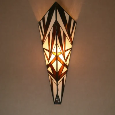 Applique murale verticale de style Art Déco en vitrail Tiffany, de forme effilée et conique. Le motif est composé de multiples triangles et formes angulaires, créant un design très dynamique et fracturé, presque abstrait. La palette de couleurs est un contraste fort et graphique entre le noir et le blanc opalescent. L'applique projette une lumière chaude vers le haut et vers le bas, accentuant son relief et son motif aiguisé.