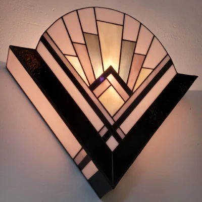 Applique murale Art Déco en Vitrail Tiffany, de forme semi-circulaire ou en éventail, symbolisant un lever de soleil. Le motif est structuré par des lignes géométriques et des rayons. La palette de couleurs se compose de verres noir opaque pour les larges bandes latérales, de blanc opalescent pour le fond principal, avec des touches subtiles de gris pâle et d'ivoire au centre. L'applique est fixée au mur et projette une lumière chaude vers le haut et sur le mur environnant, soulignant son design graphique.