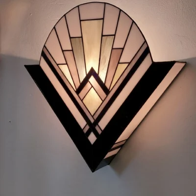 Applique murale Art Déco en Vitrail Tiffany, de forme semi-circulaire ou en éventail, symbolisant un lever de soleil. Le motif est structuré par des lignes géométriques et des rayons. La palette de couleurs se compose de verres noir opaque pour les larges bandes latérales, de blanc opalescent pour le fond principal, avec des touches subtiles de gris pâle et d'ivoire au centre. L'applique est fixée au mur et projette une lumière chaude vers le haut et sur le mur environnant, soulignant son design graphique.