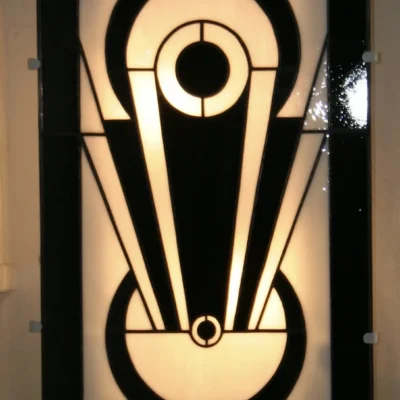 Applique murale de vitrail Art Déco, probablement. Le design est vertical et symétrique, centré sur un motif géométrique abstrait très graphique, évoquant un vase stylisé. La palette de couleurs est un contraste fort entre le noir opaque et le blanc opalescent rétroéclairé, soulignant les formes audacieuses, les cercles concentriques et les lignes évasées typiques de l'Art Déco.