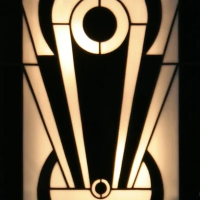 Applique murale de vitrail Art Déco, probablement. Le design est vertical et symétrique, centré sur un motif géométrique abstrait très graphique, évoquant un vase stylisé. La palette de couleurs est un contraste fort entre le noir opaque et le blanc opalescent rétroéclairé, soulignant les formes audacieuses, les cercles concentriques et les lignes évasées typiques de l'Art Déco.