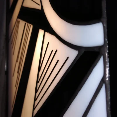 Applique murale rectangulaire verticale de style Art Déco en vitrail Tiffany. Le panneau présente un motif géométrique et cinétique asymétrique, dominé par des formes triangulaires pointues et des cercles concentriques sur le côté droit. La palette de couleurs est un contraste graphique entre le noir opaque et le blanc opalescent rétroéclairé. De fines lignes gravées (de couleur noir) soulignent l'effet de vitesse et de mouvement. La lumière douce filtre à travers le verre, créant un éclairage d'ambiance.