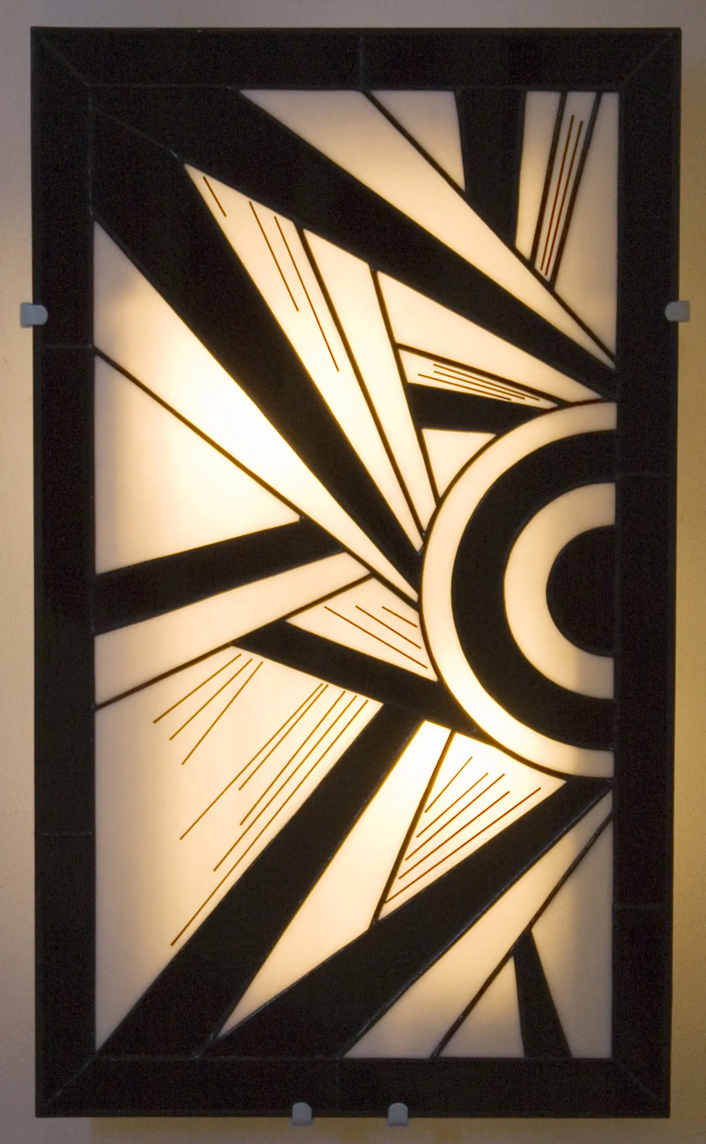 Applique murale rectangulaire verticale de style Art Déco en vitrail Tiffany. Le panneau présente un motif géométrique et cinétique asymétrique, dominé par des formes triangulaires pointues et des cercles concentriques sur le côté droit. La palette de couleurs est un contraste graphique entre le noir opaque et le blanc opalescent rétroéclairé. De fines lignes gravées (de couleur noir) soulignent l'effet de vitesse et de mouvement. La lumière douce filtre à travers le verre, créant un éclairage d'ambiance.