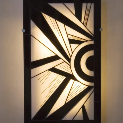 Applique murale rectangulaire verticale de style Art Déco en vitrail Tiffany. Le panneau présente un motif géométrique et cinétique asymétrique, dominé par des formes triangulaires pointues et des cercles concentriques sur le côté droit. La palette de couleurs est un contraste graphique entre le noir opaque et le blanc opalescent rétroéclairé. De fines lignes gravées (de couleur noir) soulignent l'effet de vitesse et de mouvement. La lumière douce filtre à travers le verre, créant un éclairage d'ambiance.