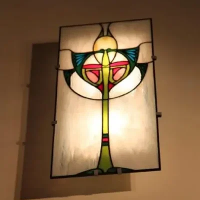 Détail d'une applique murale ou d'un panneau en vitrail de style Art Nouveau. La pièce présente un motif floral stylisé et symétrique sur fond dépoli et rétroéclairé. Les couleurs dominantes sont le jaune, le vert vif, le rouge, le rose saumon, et le bleu turquoise, encadrées par des lignes de plomb noires. Des textures de verre striées sont visibles sur les bords inférieurs du motif, évoquant des ailes ou des feuilles