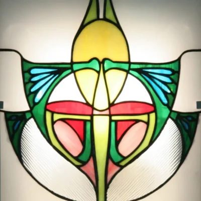 Détail d'une applique murale ou d'un panneau en vitrail de style Art Nouveau. La pièce présente un motif floral stylisé et symétrique sur fond dépoli et rétroéclairé. Les couleurs dominantes sont le jaune, le vert vif, le rouge, le rose saumon, et le bleu turquoise, encadrées par des lignes de plomb noires. Des textures de verre striées sont visibles sur les bords inférieurs du motif, évoquant des ailes ou des feuilles