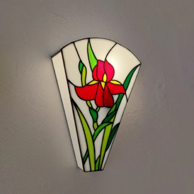 Applique murale Art Nouveau en vitrail de style Tiffany, fixée sur un mur gris texturé. L'applique a une forme évasée en éventail inversé. Elle représente une grande fleur d'iris rouge vif avec un cœur jaune, entourée de longues feuilles vertes et vert chartreuse. Les bords du vitrail sont constitués de verre blanc opaque, et l'ensemble est souligné par des lignes de soudure noires