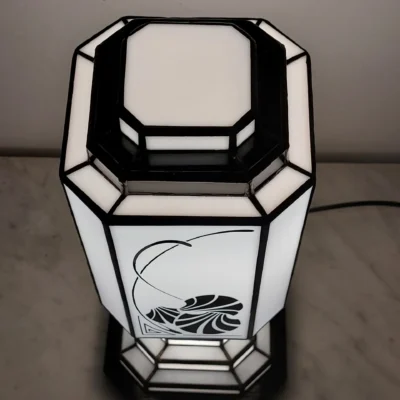 Lampe de table Art Déco en vitrail Tiffany, de forme rectangulaire et colonne, posée sur une base étagée. L'abat-jour principal est composé de panneaux de verre blanc opalescent, ornés d'un motif floral stylisé ou de coquillages en verre noir opaque, avec des lignes courbes délicates. La base et le sommet sont également en vitrail noir et blanc, avec un design géométrique et étagé qui éclaire de l'intérieur. L'ensemble diffuse une lumière douce et d'ambiance, mettant en valeur son design élégant et ses motifs inspirés de l'Art Nouveau et de l'Art Déco.