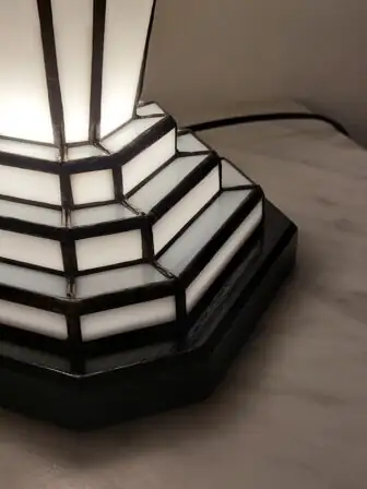 Lampe de table Art Déco en vitrail Tiffany, de forme rectangulaire et colonne, posée sur une base étagée. L'abat-jour principal est composé de panneaux de verre blanc opalescent, ornés d'un motif floral stylisé ou de coquillages en verre noir opaque, avec des lignes courbes délicates. La base et le sommet sont également en vitrail noir et blanc, avec un design géométrique et étagé qui éclaire de l'intérieur. L'ensemble diffuse une lumière douce et d'ambiance, mettant en valeur son design élégant et ses motifs inspirés de l'Art Nouveau et de l'Art Déco.