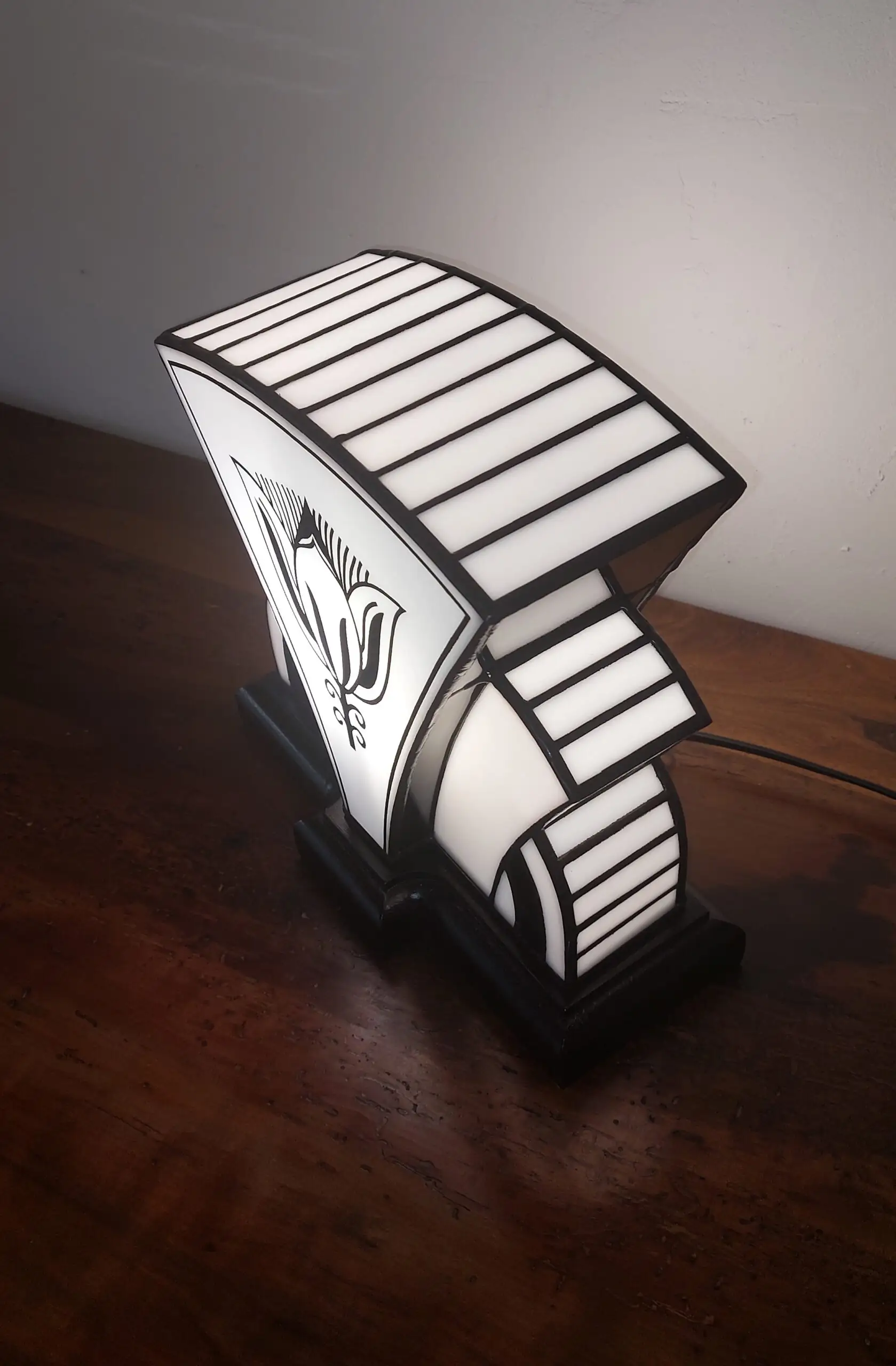 Lampe de table Art Déco de forme stylisée et symétrique, rappelant un éventail ou un "soleil levant" stylisé. Elle est composée de panneaux de vitrail en verre blanc opalescent et noir opaque. Le panneau central est orné d'un grand motif floral géométrique stylisé (fleur de lotus), réalisé en verre noir sur fond blanc. La base est noire, et l'ensemble de la lampe est illuminé de l'intérieur, projetant une lumière douce et créant un fort contraste avec les motifs noirs. Le design est un mélange élégant d'influences Art Déco et Art Nouveau.