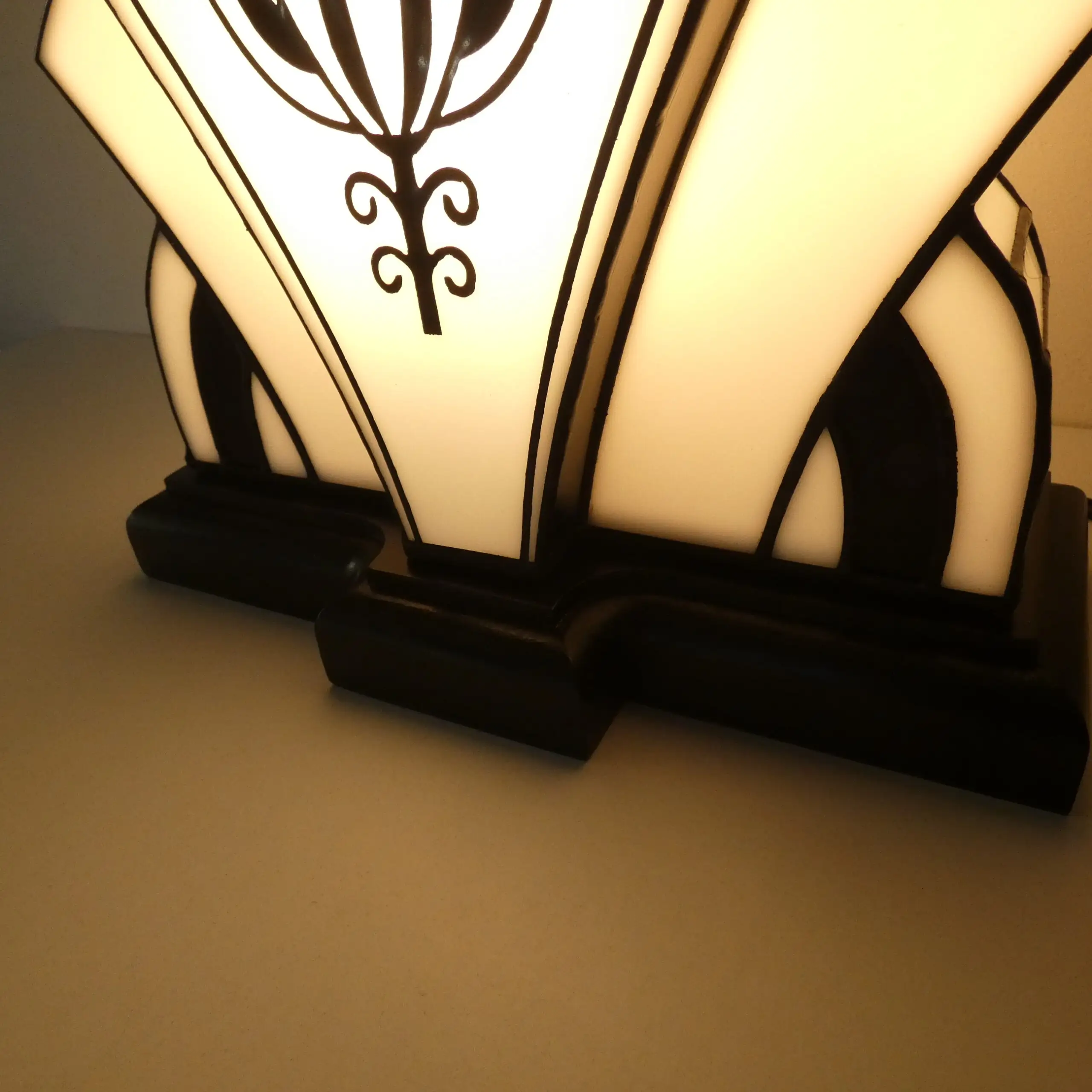 Lampe de table Art Déco de forme stylisée et symétrique, rappelant un éventail ou un "soleil levant" stylisé. Elle est composée de panneaux de vitrail en verre blanc opalescent et noir opaque. Le panneau central est orné d'un grand motif floral géométrique stylisé (fleur de lotus), réalisé en verre noir sur fond blanc. La base est noire, et l'ensemble de la lampe est illuminé de l'intérieur, projetant une lumière douce et créant un fort contraste avec les motifs noirs. Le design est un mélange élégant d'influences Art Déco et Art Nouveau.