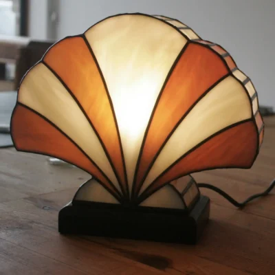 Lampe de table Art Déco en vitrail Tiffany, de forme éventail ou coquillage stylisé. Le design est composé de panneaux de verre colorés et de soudures noires, créant une structure graphique et évasée. La palette de couleurs alterne des sections de blanc opalescent avec des nuances chaudes d'orange, de miel et d'ambre, rappelant un coucher de soleil ou des couleurs automnales. La base est sombre (noir ou bois foncé), et la lampe est illuminée de l'intérieur, diffusant une lumière chaude et invitante.