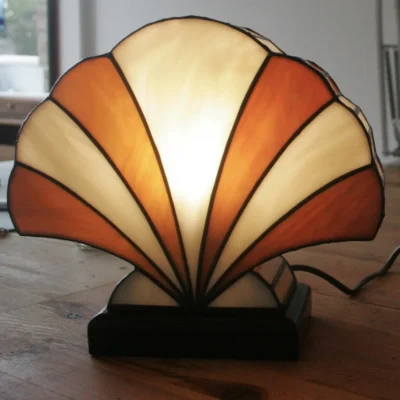 Lampe de table Art Déco en vitrail Tiffany, de forme éventail ou coquillage stylisé. Le design est composé de panneaux de verre colorés et de soudures noires, créant une structure graphique et évasée. La palette de couleurs alterne des sections de blanc opalescent avec des nuances chaudes d'orange, de miel et d'ambre, rappelant un coucher de soleil ou des couleurs automnales. La base est sombre (noir ou bois foncé), et la lampe est illuminée de l'intérieur, diffusant une lumière chaude et invitante.