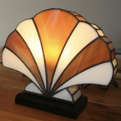 Lampe de table Art Déco en vitrail Tiffany, de forme éventail ou coquillage stylisé. Le design est composé de panneaux de verre colorés et de soudures noires, créant une structure graphique et évasée. La palette de couleurs alterne des sections de blanc opalescent avec des nuances chaudes d'orange, de miel et d'ambre, rappelant un coucher de soleil ou des couleurs automnales. La base est sombre (noir ou bois foncé), et la lampe est illuminée de l'intérieur, diffusant une lumière chaude et invitante.