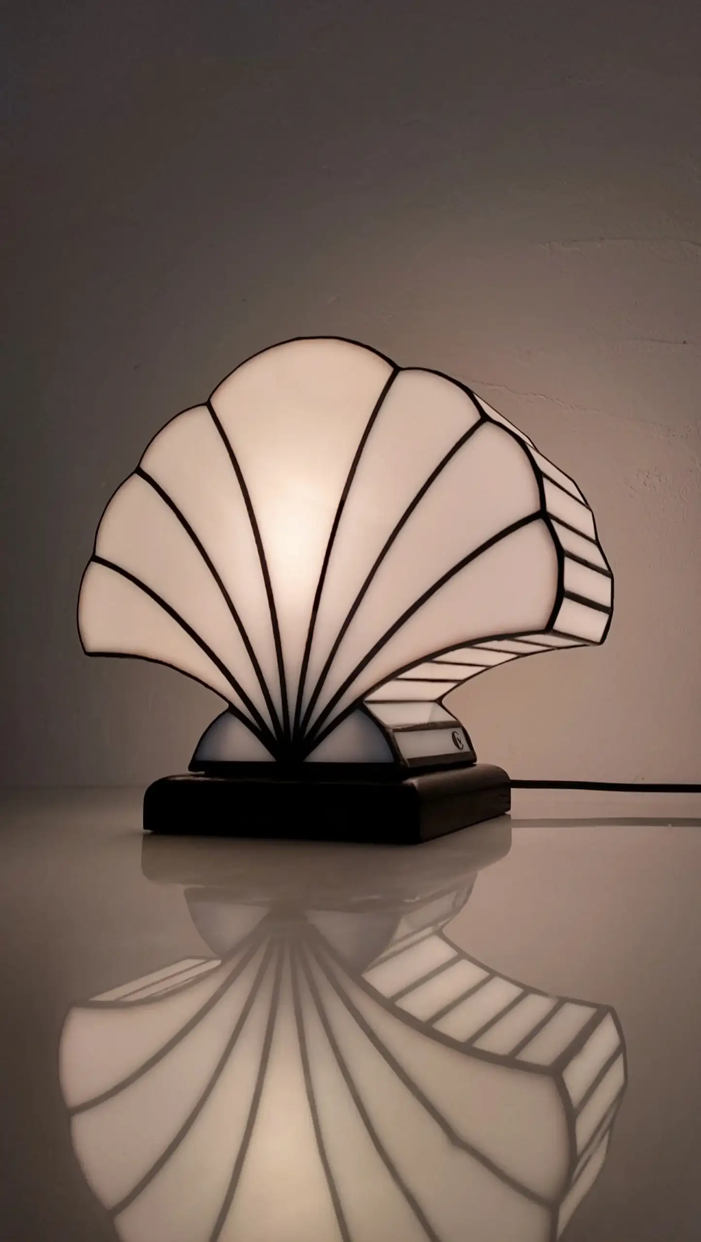 Lampe de table Art Déco en vitrail Tiffany, de forme coquillage stylisé. Le design est composé de panneaux de verre blanc opalescent et de soudures noires, créant une structure graphique et évasée. La base est noire, et la lampe est illuminée de l'intérieur, diffusant une lumière douce et uniforme qui met en valeur la forme et les lignes noires. Son reflet clair est visible sur la surface brillante sous la lampe, accentuant son élégance classique inspirée des années 1920-1930.